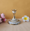 A single candle stand with a wide, circular base made of brass and inlaid with mother-of-pearl or similar iridescent material. The stand has a slender, tapering stem and is shown on a light-colored surface with two flowers on either side.