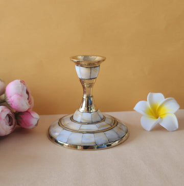 A single candle stand with a wide, circular base made of brass and inlaid with mother-of-pearl or similar iridescent material. The stand has a slender, tapering stem and is shown on a light-colored surface with two flowers on either side.