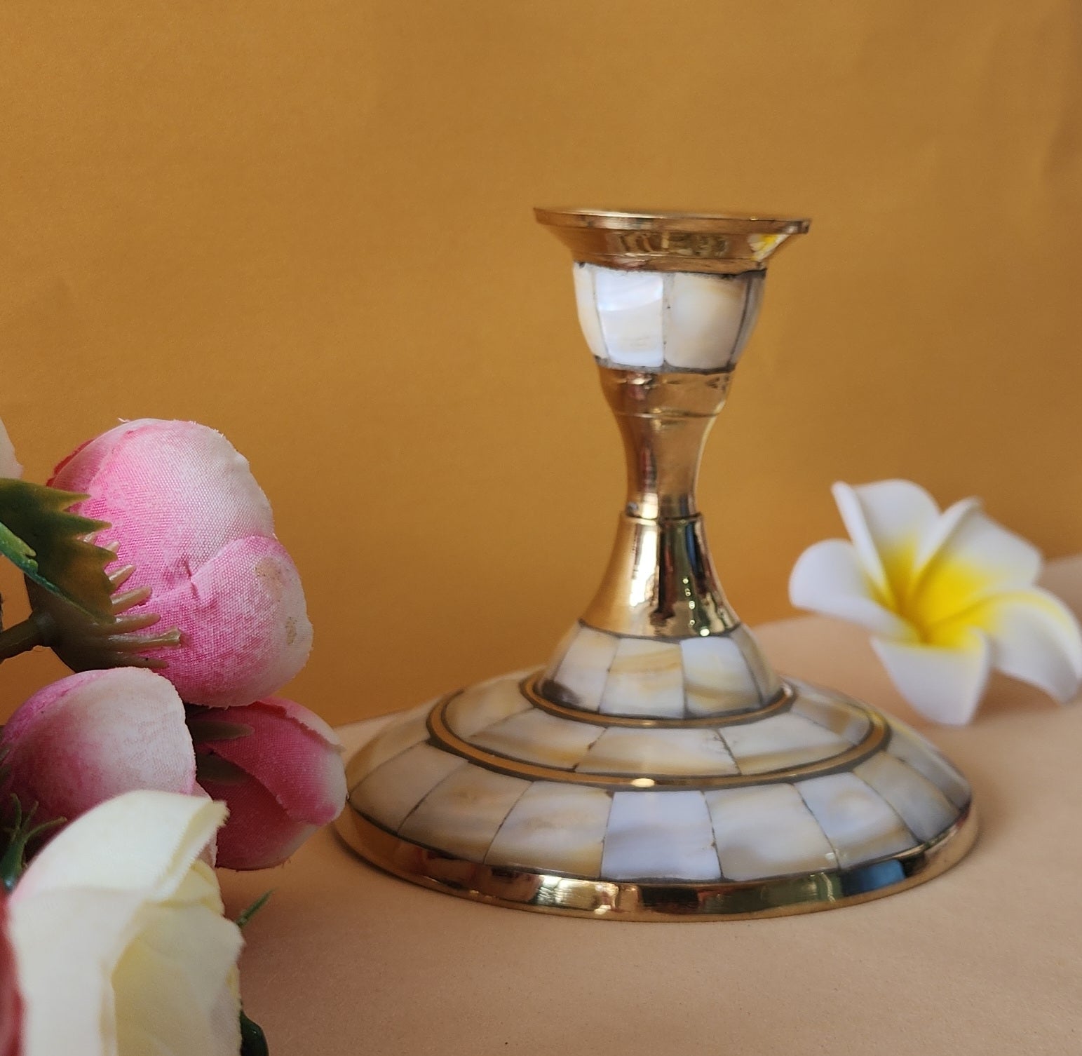 A single candle stand with a wide, circular base made of brass and inlaid with mother-of-pearl or a similar iridescent material. The stand has a tapering stem and is placed on a light-colored surface with two flowers on either side.