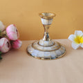 A single candle stand with a wide, circular base made of brass and inlaid with mother-of-pearl or a similar iridescent material. The stand has a tapering stem and is placed on a light-colored surface with two flowers on either side.