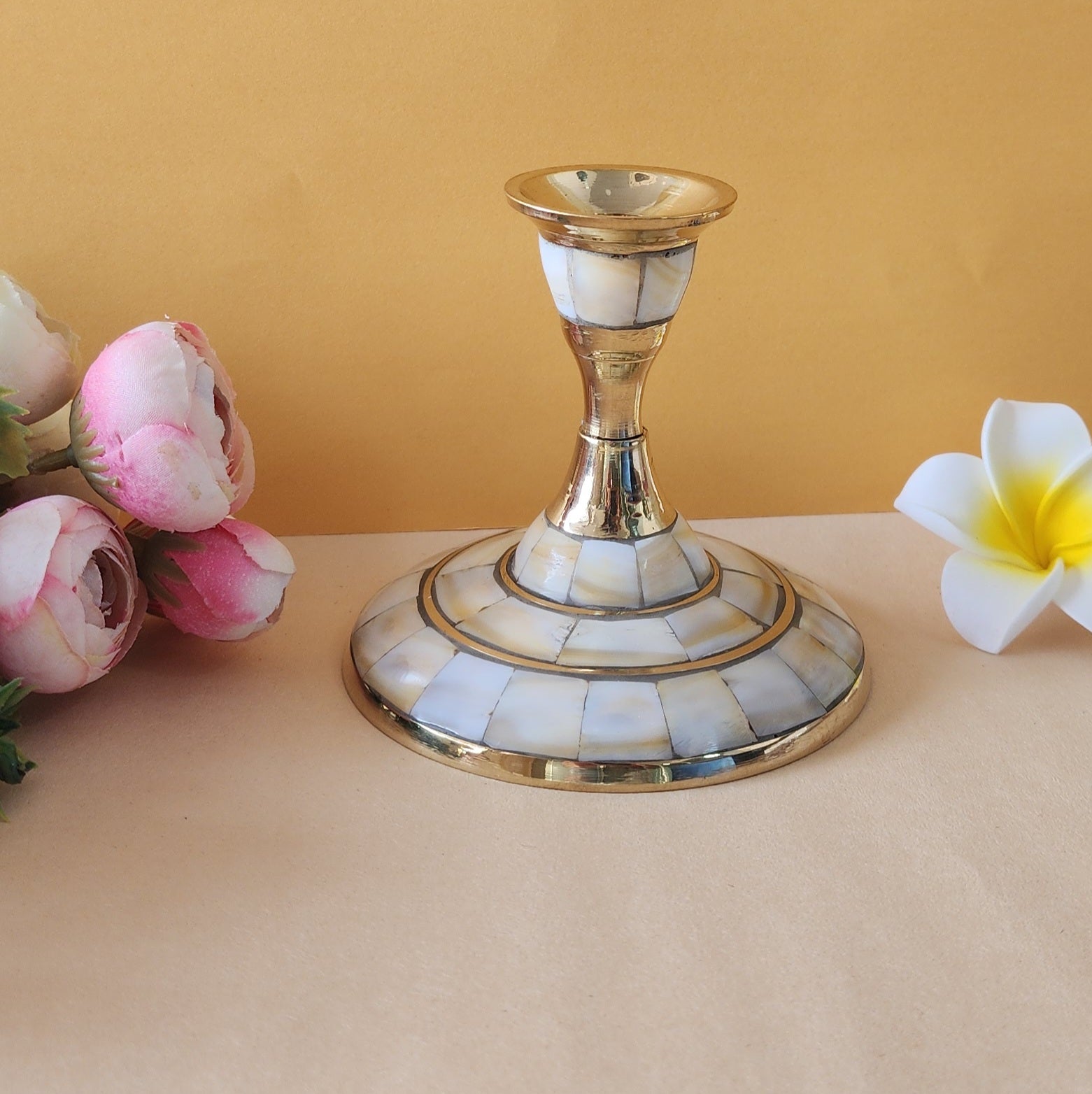 A single candle stand with a wide, circular base made of brass and inlaid with mother-of-pearl or a similar iridescent material. The stand has a tapering stem and is placed on a light-colored surface with two flowers on either side.