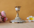 A single candle stand with a wide, circular base made of brass and inlaid with mother-of-pearl or a similar iridescent material. The stand has a tapering stem and is placed on a light-colored surface with two flowers on either side.