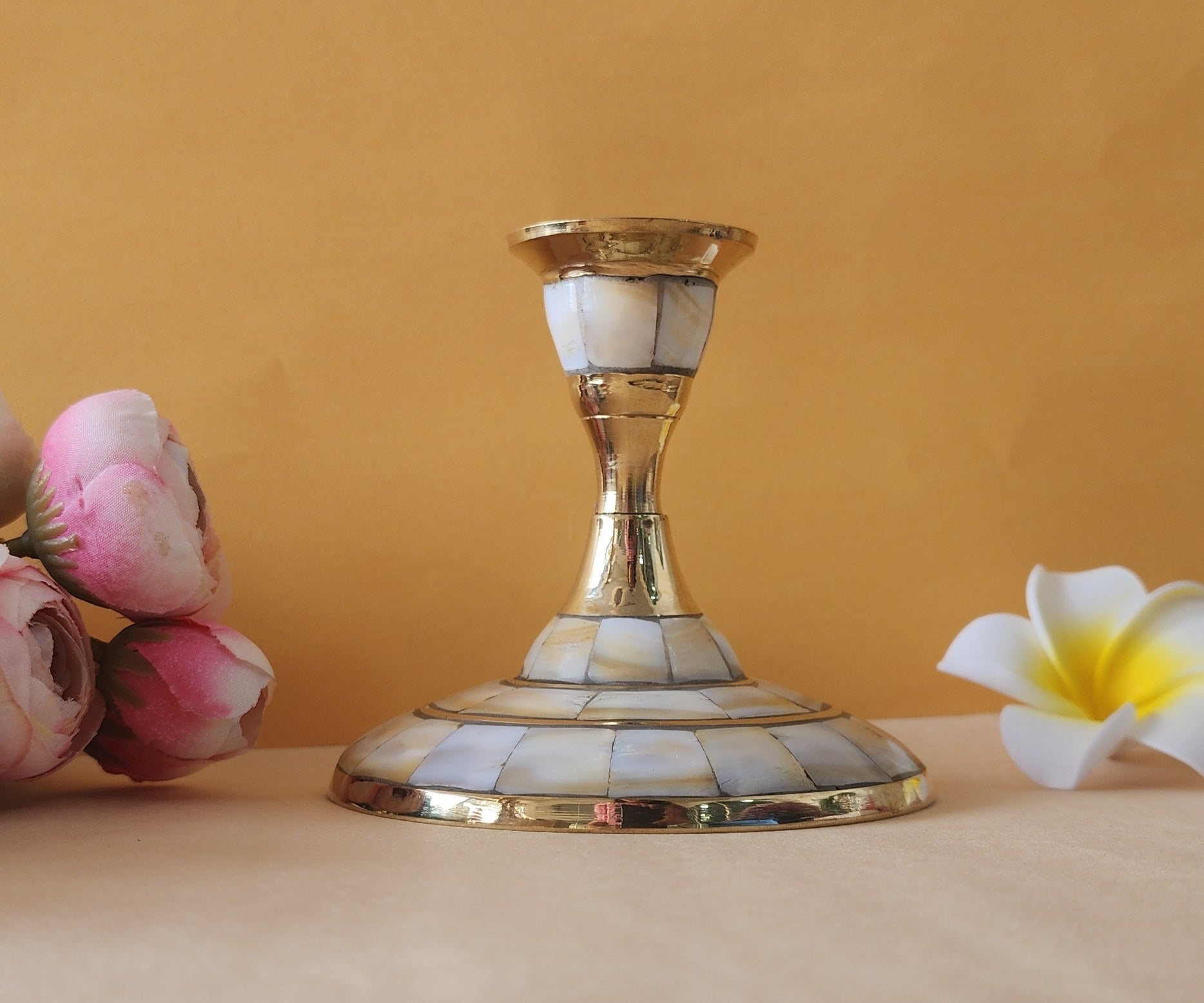 A single candle stand with a wide, circular base made of brass and inlaid with mother-of-pearl or a similar iridescent material. The stand has a tapering stem and is placed on a light-colored surface with two flowers on either side.