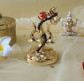 A golden decorative idol featuring a Trishul (trident) entwined with a small drum (Damru) and adorned with the sacred Om symbol, resting on a textured base. In the background, out of focus, a white frangipani flower and another golden idol are visible.