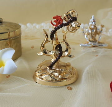 A golden decorative idol featuring a Trishul (trident) entwined with a small drum (Damru) and adorned with the sacred Om symbol, resting on a textured base. In the background, out of focus, a white frangipani flower and another golden idol are visible.