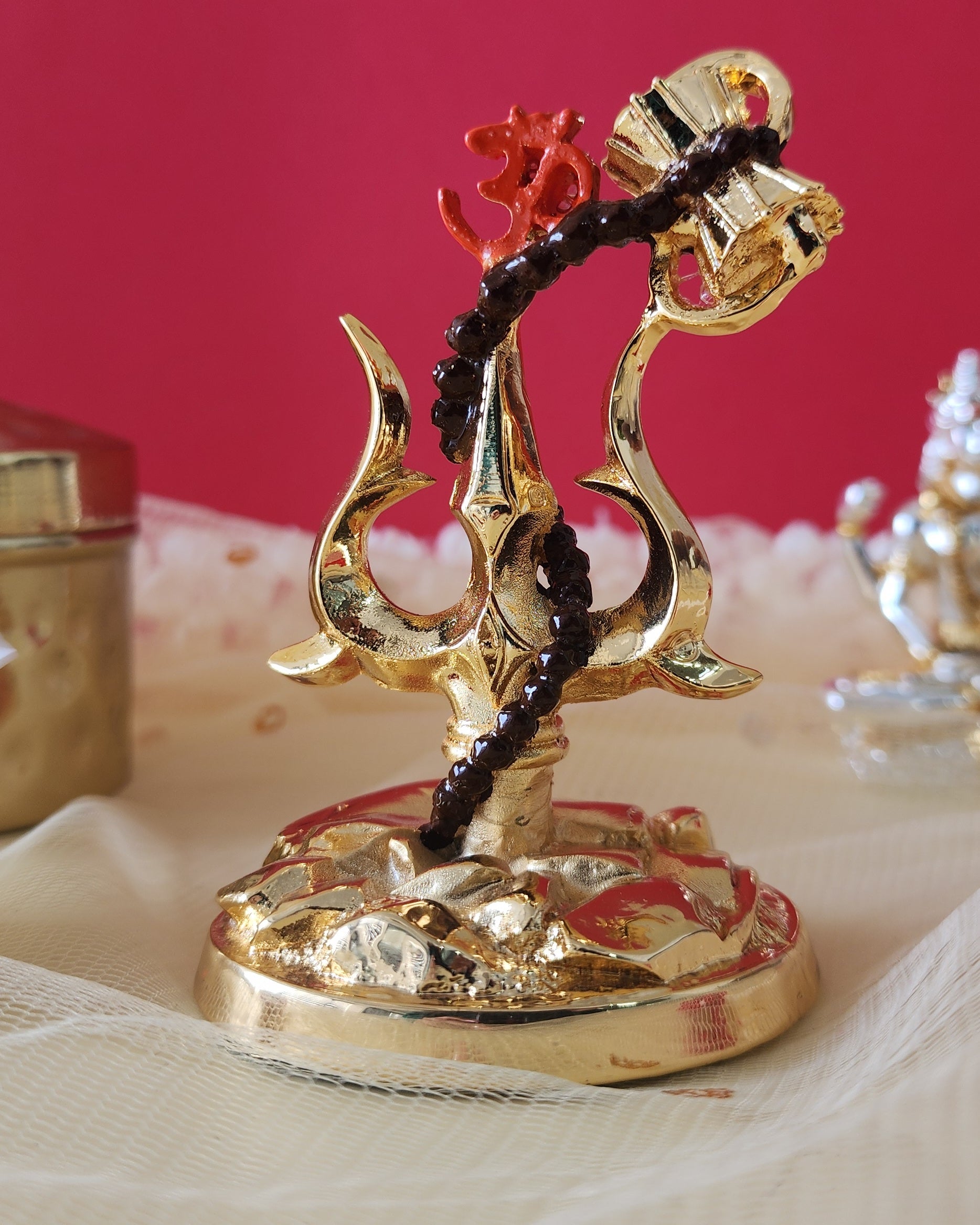 A gold-toned decorative showpiece featuring Lord Shiva's Trishul (trident) with an attached Damru (small drum) and a string of dark beads wrapped around it, set against a warm, reddish background with a lighter fabric surface below.
