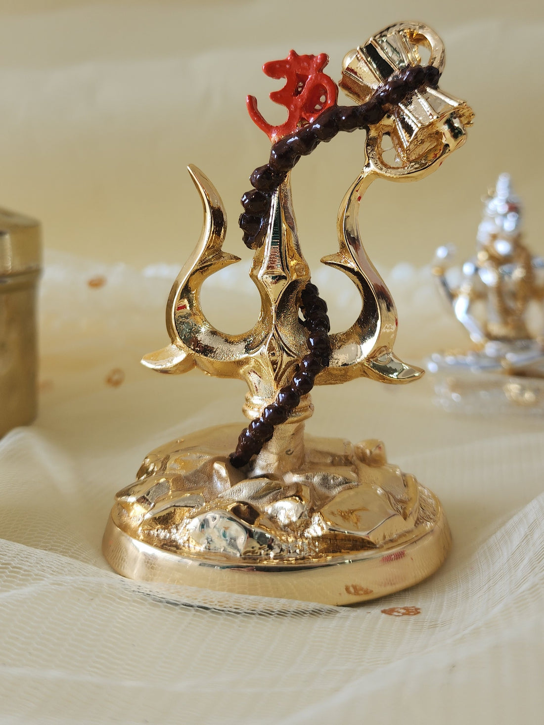 A golden decorative idol featuring a Trishul (trident) entwined with a small drum (Damru) and adorned with the sacred Om symbol, resting on a textured base. In the background, out of focus, a white frangipani flower and another golden idol are visible.