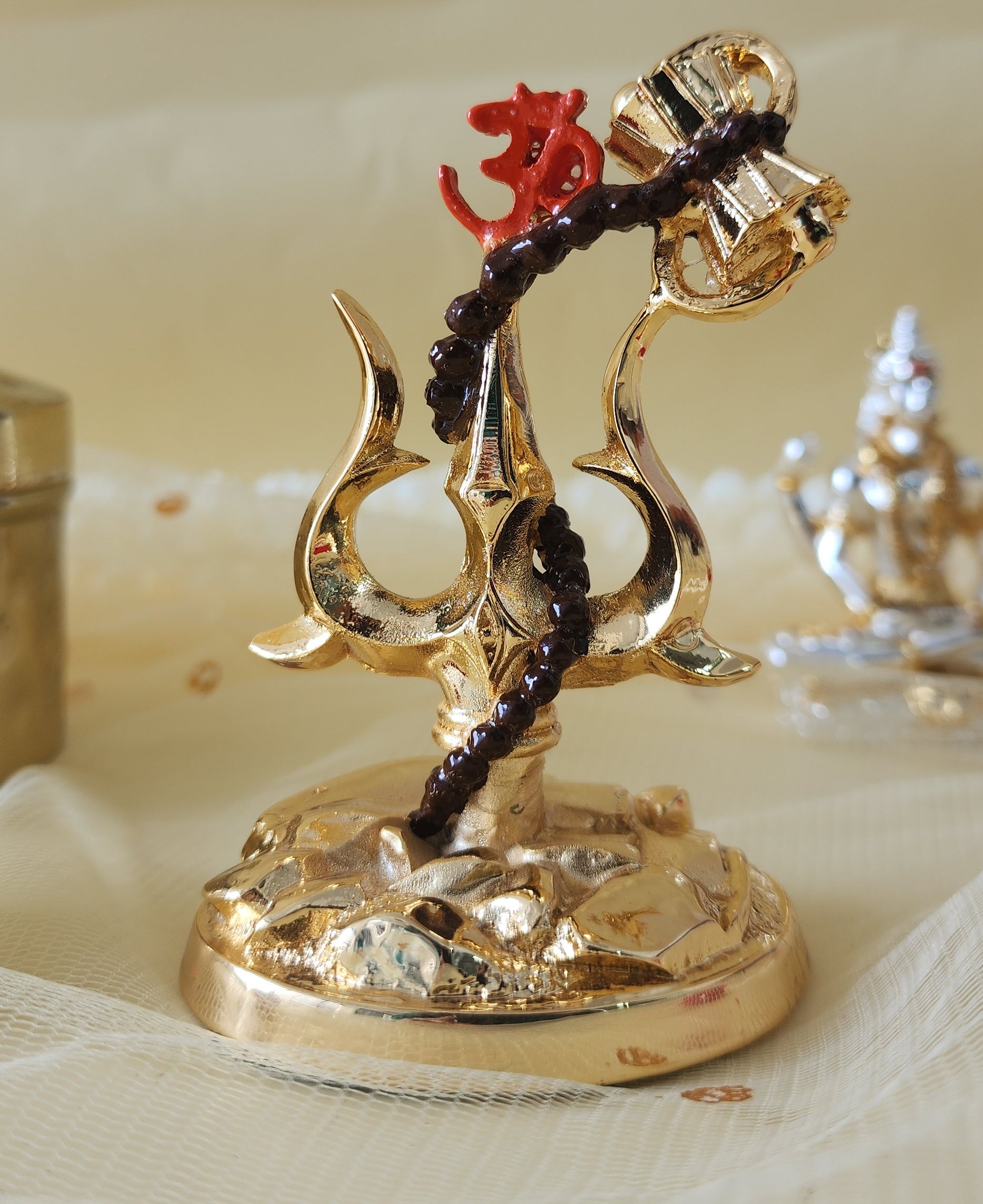 A golden Trishul, a traditional Hindu religious symbol, adorned with a small red Om symbol at its top and a miniature damru (drum) attached by a brown string, standing on an ornate golden base.