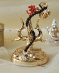 A golden religious idol featuring Lord Shiva's Trishul (trident) entwined with a Damru (drum) and topped with a red Om symbol, placed on a decorative base against a blurred, warm-toned background.