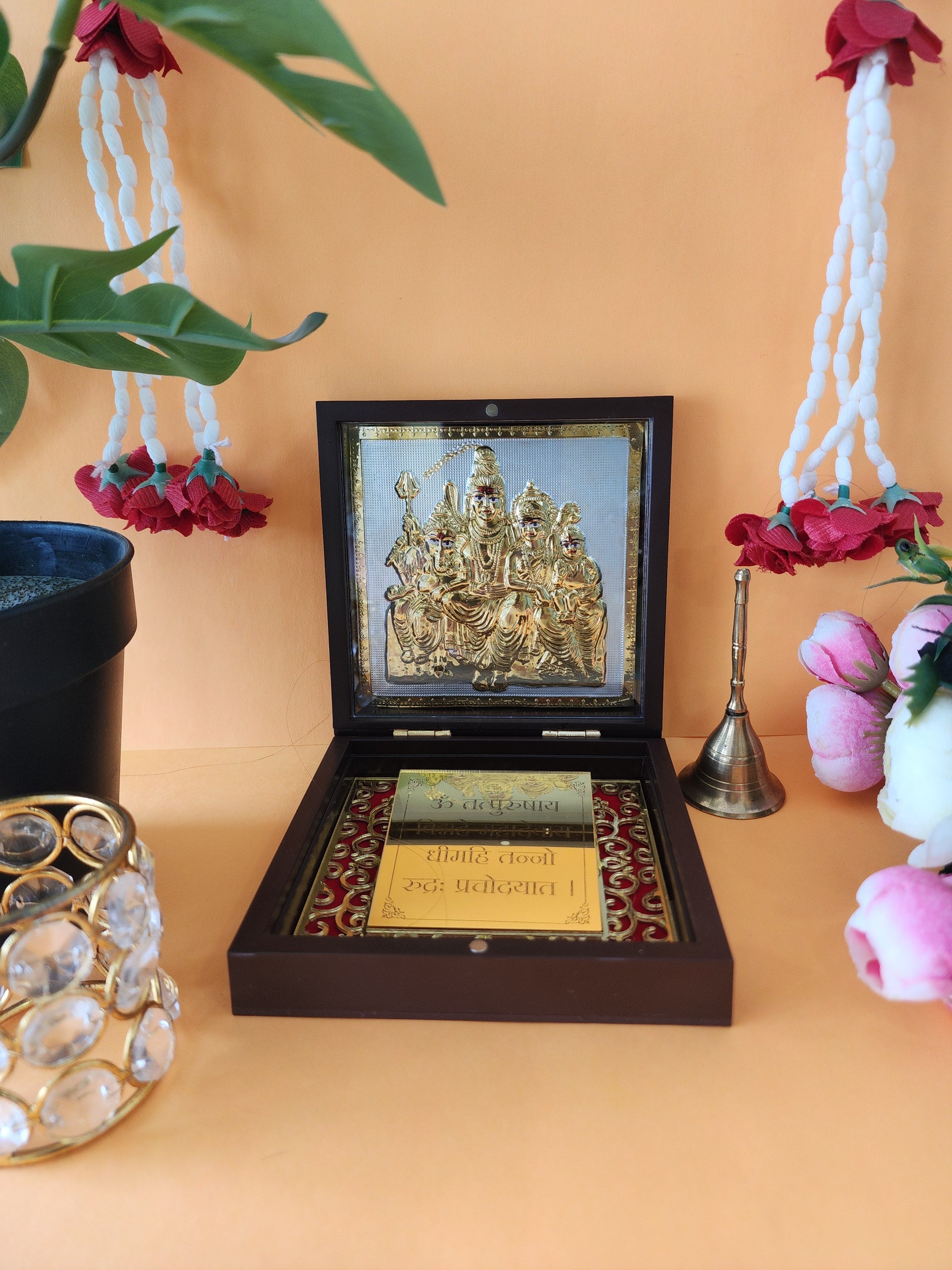 An open dark brown wooden box containing a gold-colored idol set of the Shiv Parivar (Lord Shiva, Goddess Parvati, Lord Ganesha, and Lord Kartikeya) in an intricate design. The inside of the box is adorned with red and gold patterns. The setting includes hanging white and red flower garlands, a plant in a black pot, a brass bell, and pink and white decorative flowers in the background, creating a serene, spiritual ambiance.