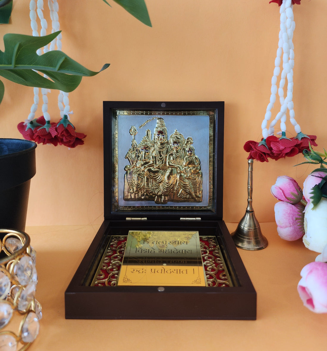 A brown wooden box, presented open, showcasing a golden, intricately designed idol or framed image of the Shiva Parivar (Lord Shiva, Goddess Parvati, Lord Ganesha, and Lord Kartikeya) placed on a golden and red velvet base. Below the idol, a golden plate with text in Devanagari script, possibly a mantra or prayer, is visible. Decorative elements like white and red floral garlands hang on either side, and a small brass bell is placed to the right of the open box.