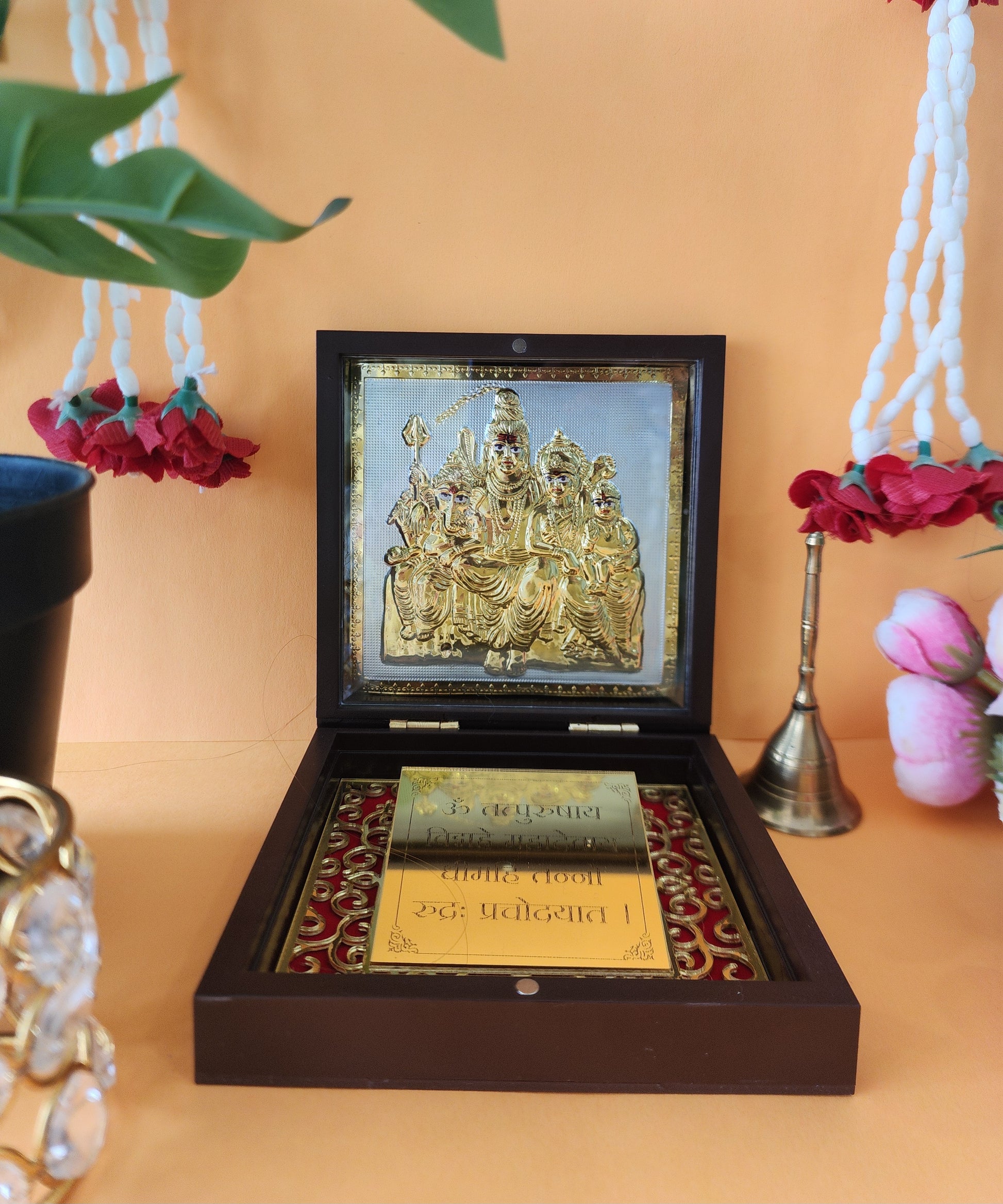 An open, dark wooden box revealing a golden framed image or idol of the Shiva Parivar (Lord Shiva, Goddess Parvati, and their children Ganesha and Kartikeya) on a textured background. Below the framed deity are two small, golden plaques with Sanskrit or Devanagari script. The box sits on a peach-colored surface, with glimpses of white and red flower garlands and a small brass bell in the background, suggesting a spiritual or devotional setting.