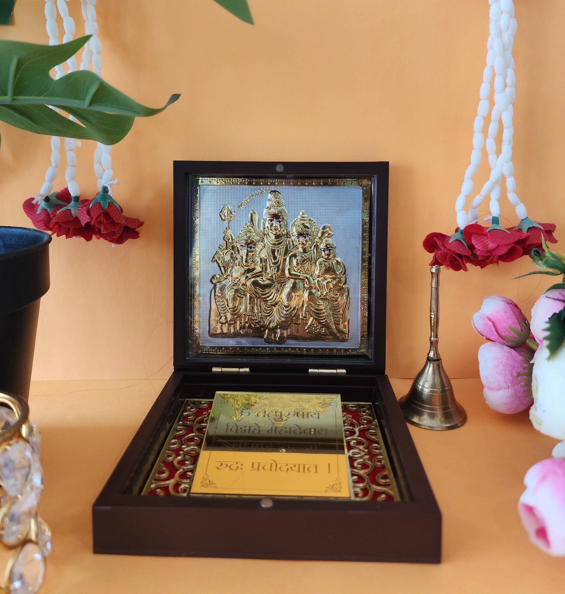 A close-up of a brown wooden box containing a gold-plated framed image of Shiva Parivar (Lord Shiva, Goddess Parvati, Lord Ganesha, and Lord Kartikeya), with religious text inscribed below, and sacred objects like a bell and white floral garlands visible in the background.