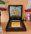 A dark wooden box, designed as a Shiva Parivar Pocket Temple, stands open, revealing a gold-plated relief of Lord Shiva, Goddess Parvati, Lord Ganesha, and Lord Kartikeya on one side, and a reflective golden panel with a Sanskrit mantra on the other. The box is set against an orange-brown background, with religious items like a bell and floral garlands visible around it, suggesting a devotional setting.
