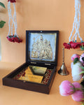 A small, open dark brown wooden box containing a gold-framed depiction of the Shiva Parivar (Lord Shiva, Goddess Parvati, Lord Ganesha, and Lord Kartikeya), with a golden bell beside it and decorative elements like flower garlands and pink roses in the background.