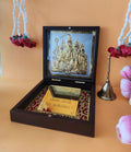 A wooden box with an open lid, revealing a gold-plated idol of Shiva Parivar (Lord Shiva, Goddess Parvati, Lord Ganesha, and Lord Kartikeya) placed on a metallic mesh background inside the lid. The lower part of the box contains a gold-colored plate with Sanskrit text and decorative border patterns, and a brass bell is visible to the right. This setup is often referred to as a
