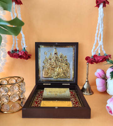 A brown wooden box, presented open, showcasing a golden, intricately designed idol or framed image of the Shiva Parivar (Lord Shiva, Goddess Parvati, Lord Ganesha, and Lord Kartikeya) placed on a golden and red velvet base. Below the idol, a golden plate with text in Devanagari script, possibly a mantra or prayer, is visible. Decorative elements like white and red floral garlands hang on either side, and a small brass bell is placed to the right of the open box.