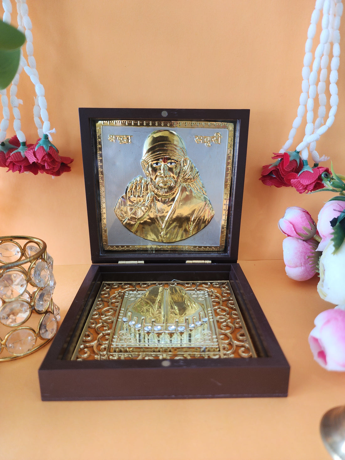 An open wooden box containing a gold-plated Shirdi Sai Baba idol and sacred Charan Paduka, placed on a light-colored surface with decorative flowers and a crystal votive holder in the background.