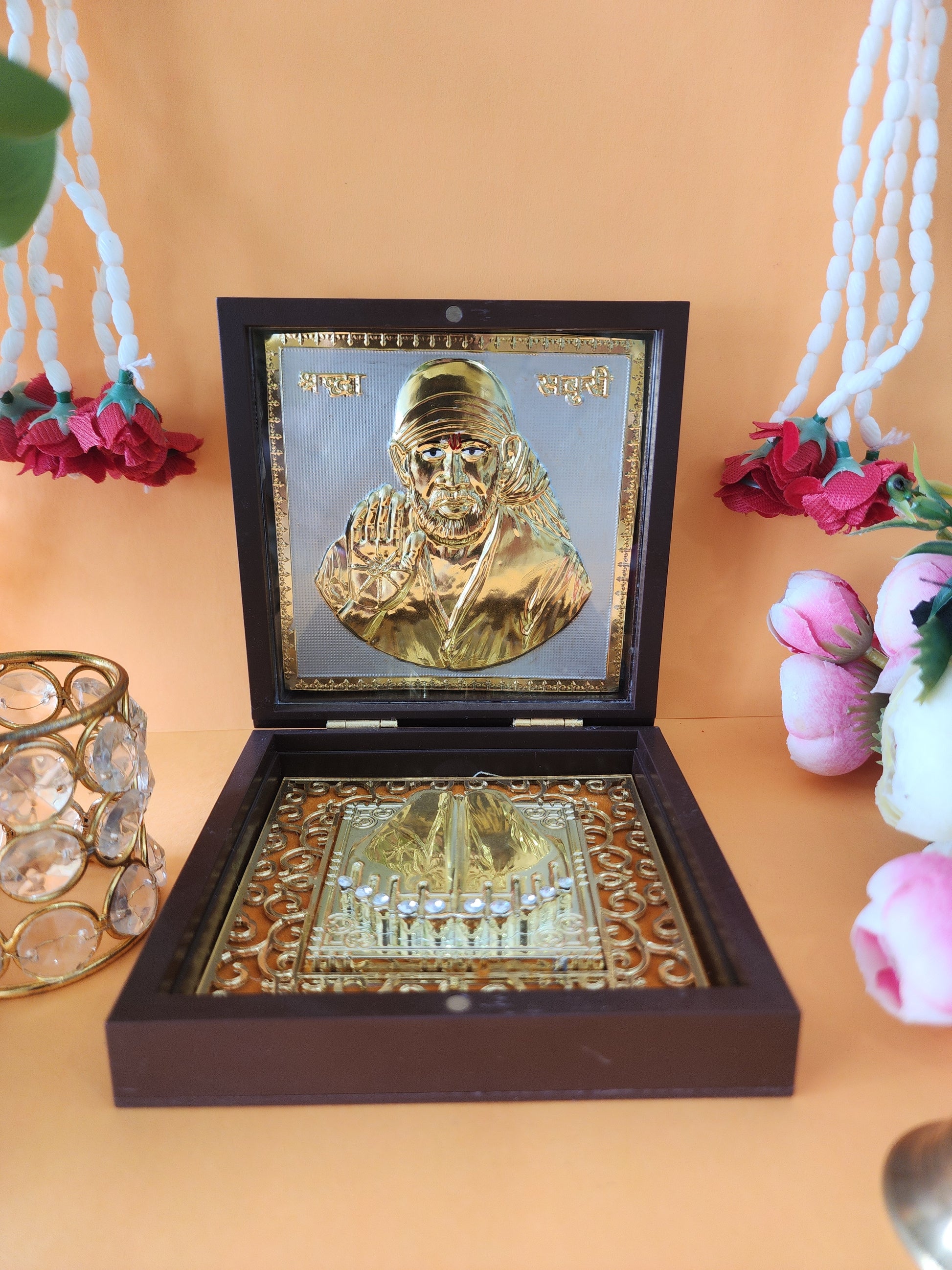 An open wooden box containing a gold-plated Shirdi Sai Baba idol and sacred Charan Paduka, placed on a light-colored surface with decorative flowers and a crystal votive holder in the background.