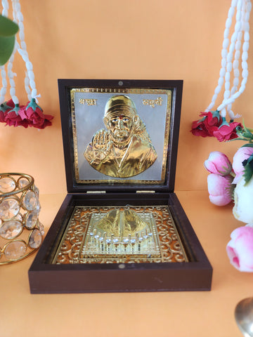 An open wooden box containing a gold-plated Shirdi Sai Baba idol and sacred Charan Paduka, placed on a light-colored surface with decorative flowers and a crystal votive holder in the background.