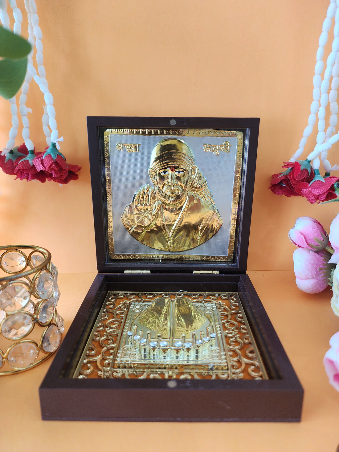 An open wooden box containing a gold-plated Shirdi Sai Baba idol and sacred Charan Paduka, placed on a light-colored surface with decorative flowers and a crystal votive holder in the background.