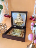 A golden-plated devotional frame featuring an image of Sai Baba with his hands raised in blessing on the top half, and golden Charan Paduka (feet impressions) on the bottom half, all housed within an open wooden box. The box is set on a light background with decorative elements like flower garlands and a small bell visible in the foreground and background.