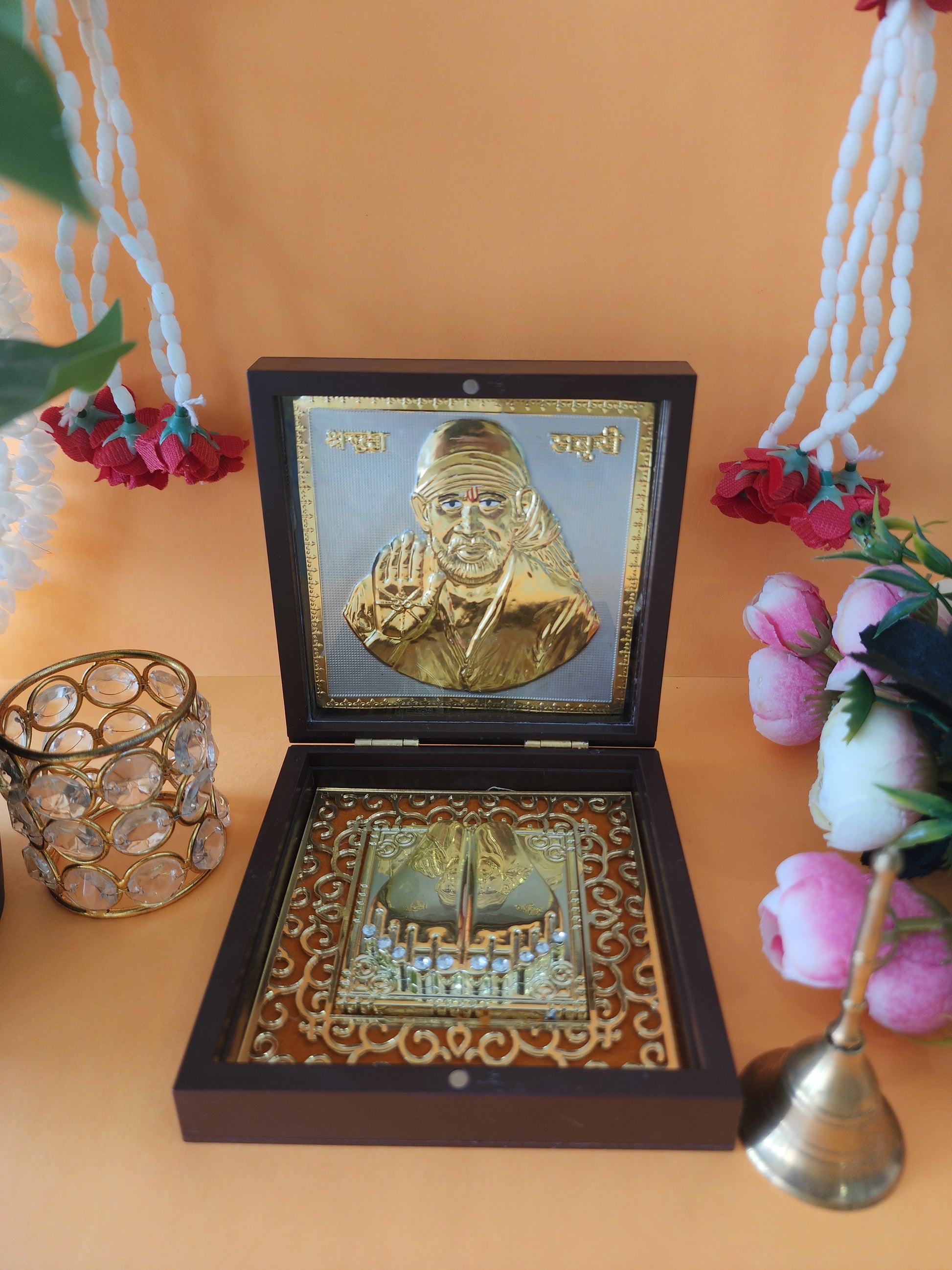 A golden-colored Sai Baba photo frame and Charan Paduka (footprints) set displayed in an open wooden box, accompanied by a decorative glass candle holder, white and red flower garlands, and a small brass bell, all arranged on an orange surface.