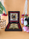 A richly adorned Ram Lalla Pocket Temple, featuring a framed image of Lord Ram Lalla in a black and gold frame, placed above a gold-plated Charan Paduka (divine footprints) within a dark wooden box lined with red velvet. The box is presented on a light orange surface, flanked by a decorative gold crystal holder on the left and pink artificial flowers and white prayer beads on the right, all against a warm background.