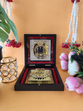 A Ram Lalla Pocket Temple open in a dark wooden box, featuring a framed image of a deity above golden Charan Paduka (footprints of a deity) on a red velvet lining, with a crystal-encrusted votive candle holder and pink roses in the background.