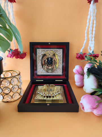 A Ram Lalla Pocket Temple open in a dark wooden box, featuring a framed image of a deity above golden Charan Paduka (footprints of a deity) on a red velvet lining, with a crystal-encrusted votive candle holder and pink roses in the background.