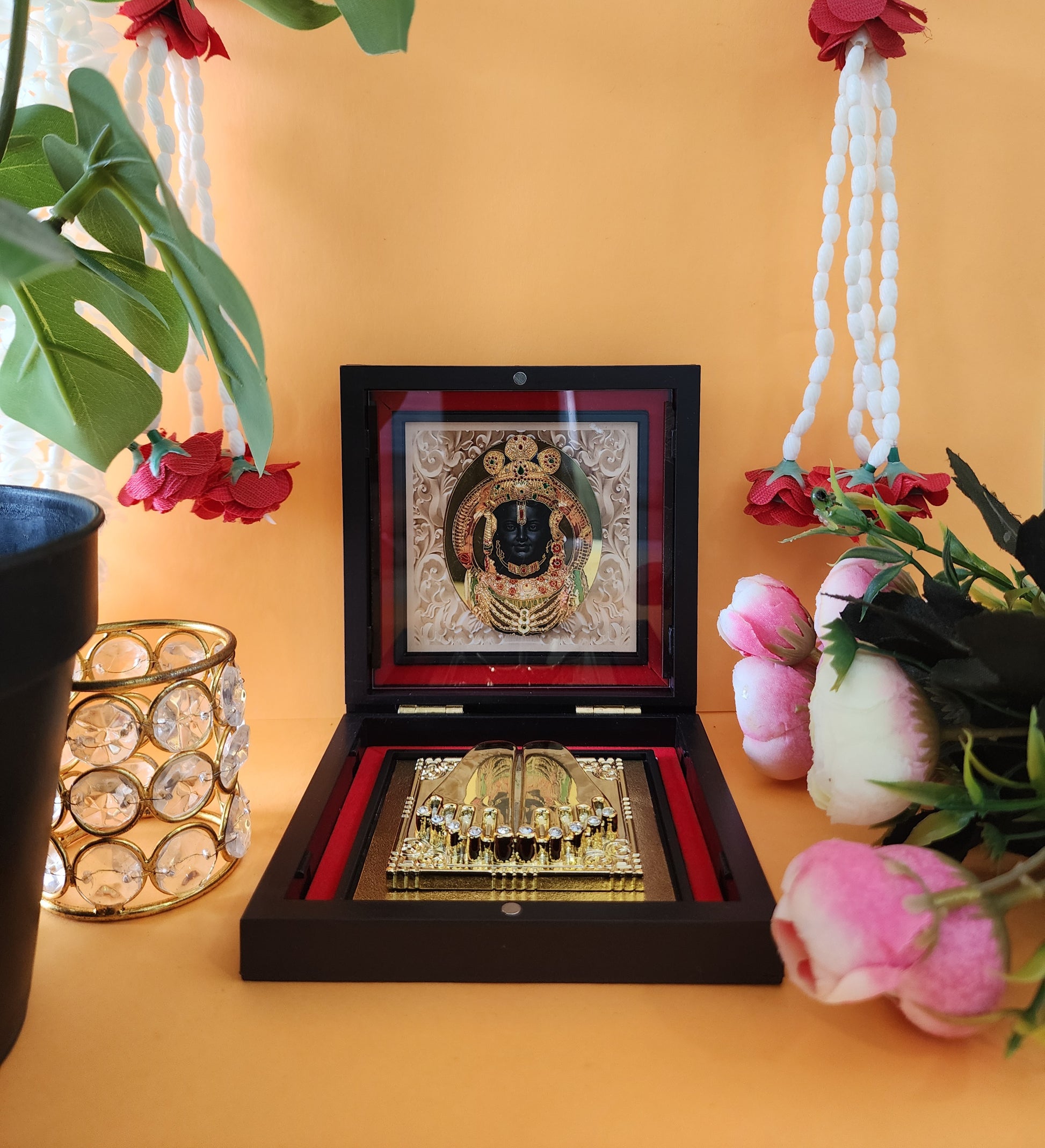 A beautifully presented Ram Lalla Pocket Temple, featuring a framed image of Lord Ram and Charan Paduka, housed in a decorative wooden box, surrounded by flowers, garlands, and a sparkling candle holder, set against an orange backdrop.