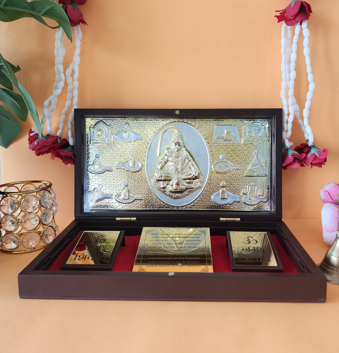 A gold-plated Lord Shiva Pocket Temple, featuring a central idol surrounded by smaller deities or symbols, displayed in an open wooden box with a red velvet interior and two additional gold-colored plaques with "Om" symbols.