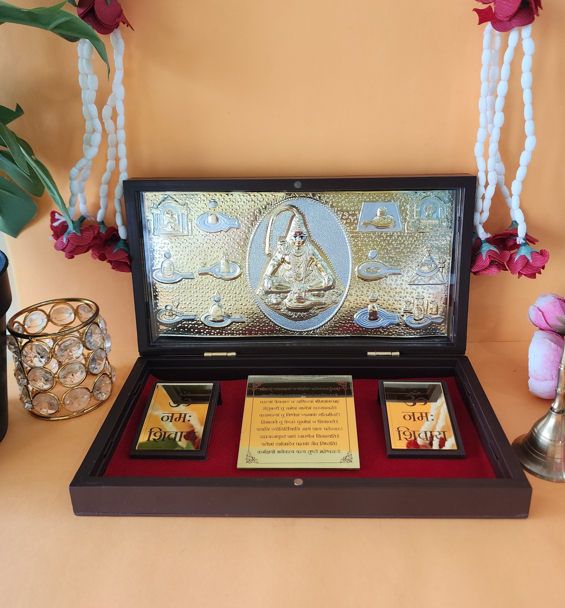 A close-up of a decorative Lord Shiva pocket temple or portable shrine, featuring a gold-plated relief of Lord Shiva surrounded by sacred symbols and perhaps the 12 Jyotirlingas, encased in a dark wooden box with a red velvet interior. The words "Om Namah Shivaya" are visibly inscribed inside the box on two plaques. A brass bell and a crystal-adorned holder are visible on either side of the box in the foreground.