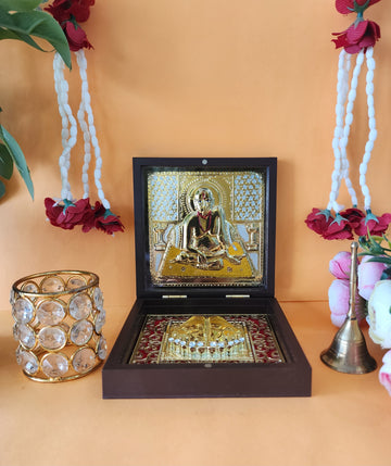 A wooden Swami Samarth Pocket Temple box open to reveal a gold-plated Swami Samarth image and Charan Paduka, flanked by decorative items including a crystal tea light holder and a brass bell, against a warm orange background with floral garlands.