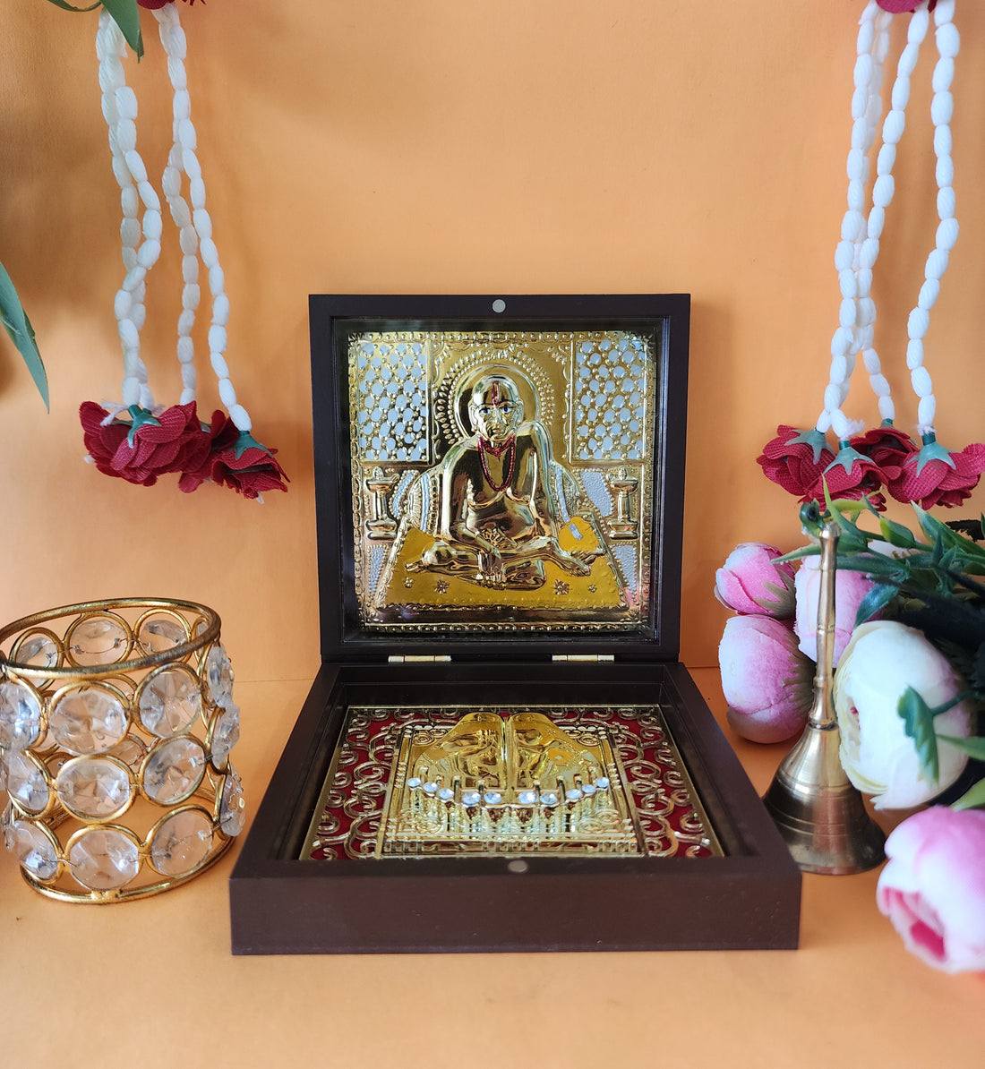 A wooden Swami Samarth Pocket Temple box open to reveal a gold-plated Swami Samarth image and Charan Paduka, flanked by decorative items including a crystal tea light holder and a brass bell, against a warm orange background with floral garlands.