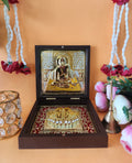 An open wooden box displaying a gold-plated image of Swami Samarth on the lid and gold-plated Charan Padukas on the base, surrounded by white beaded garlands with red flowers and a small golden bell.