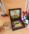 A gold-plated Swami Samarth pocket temple, open to reveal an image of Swami Samarth and Charan Paduka (divine footprints) inside a decorative wooden box, surrounded by traditional Hindu prayer items like a decorative candle holder and flower garlands.
