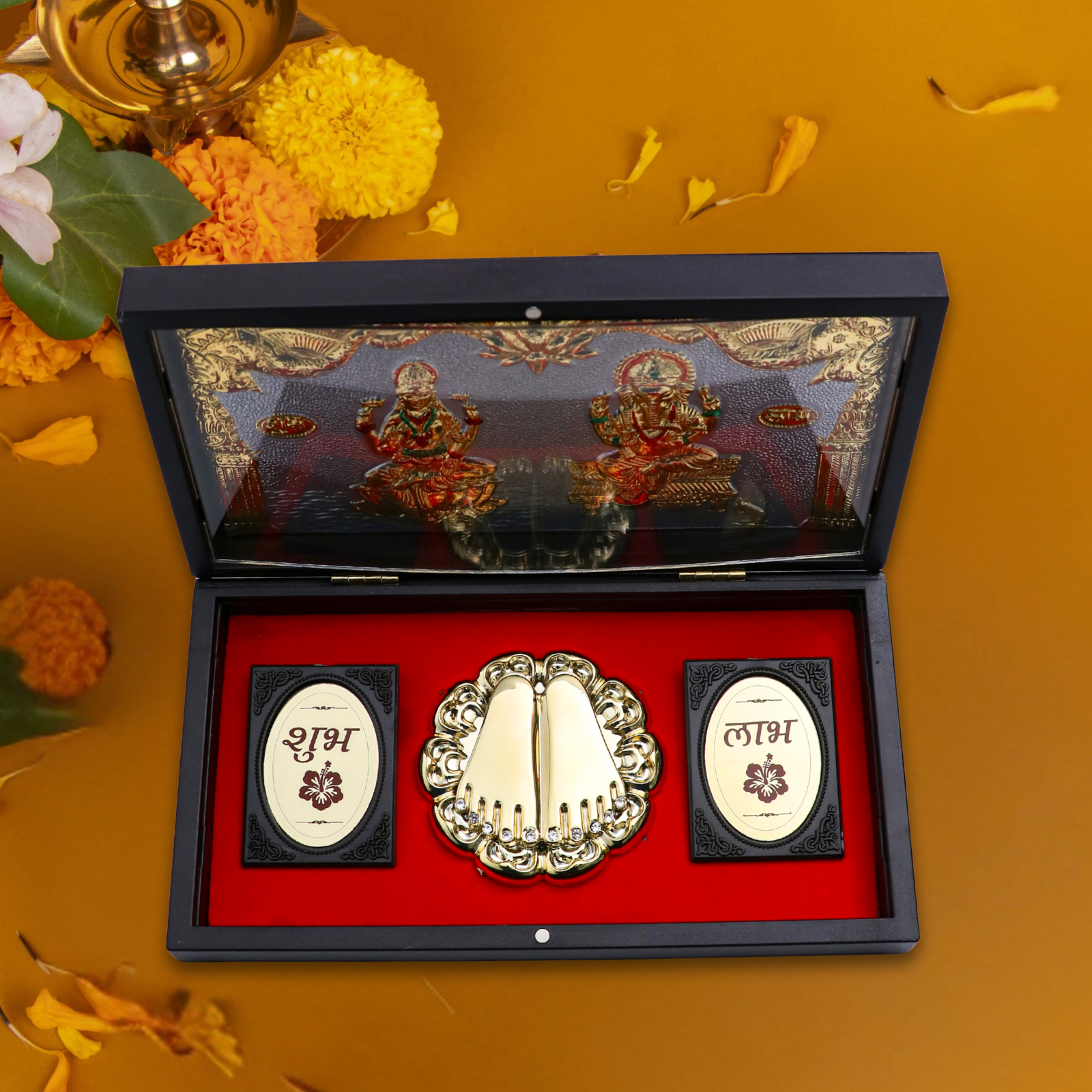 A black gift box with a red velvet interior containing a golden pair of Lakshmi Charan Paduka (sacred footprints) in the center, flanked by two golden plaques with (Shubh) and (Labh) written on them. The box lid, partially visible, depicts golden embossed images of Lord Ganesha and Goddess Lakshmi.