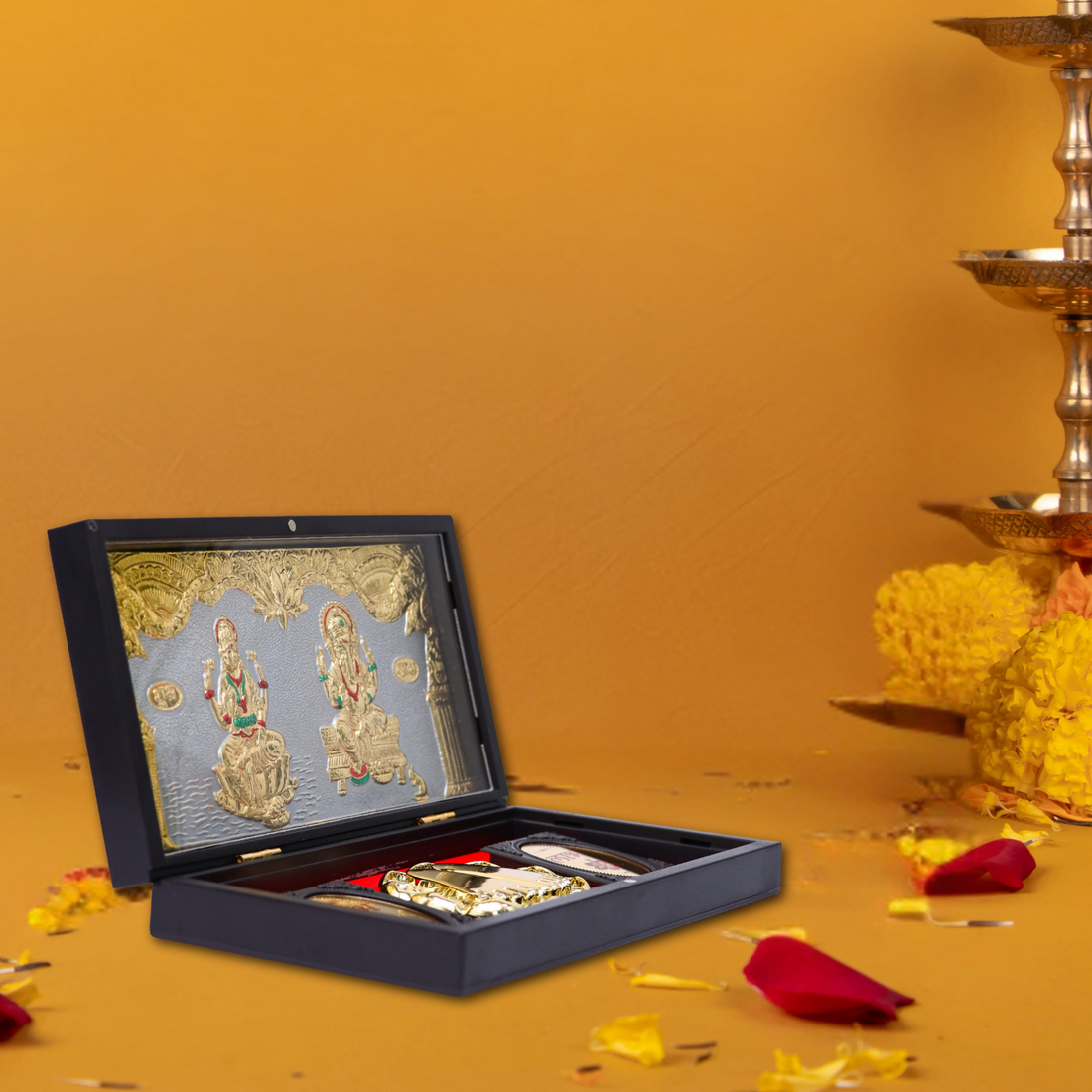 A detailed image showing a traditional Hindu puja set. The centerpiece is an open black display box with a red velvet interior, showcasing silver and gold-toned idols of Goddess Lakshmi and Lord Ganesha on a decorative backdrop. Below them are two gold-colored Laxmi Charan Paduka (footprints of Goddess Lakshmi) and a gold coin, symbolizing wealth and blessings. In the background, out of focus, are golden lamps, flower garlands, and sweets, suggesting a festive or devotional setting like Diwali celebrations.