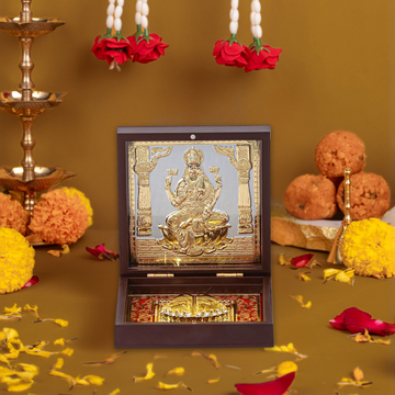 An open, dark wooden pooja box featuring a golden relief of Goddess Laxmi on the inside of the lid. The lower compartment contains a set of Golden Laxmi Charan Paduka (sacred footprints), all set against a red decorative background. The box is placed on a festive surface with marigold and rose petals, and traditional sweets and diyas are visible in the background.