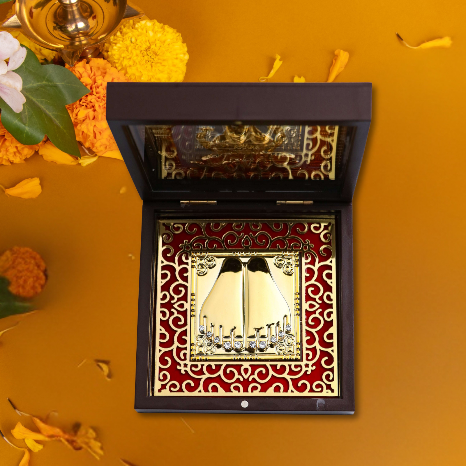 An open, dark wooden box containing a gold-plated Goddess Laxmi Paduka (sacred footprints) set against a red, ornate background. The box is placed on an orange surface with scattered flower petals, and the lid has a reflective, gold-patterned inner surface.