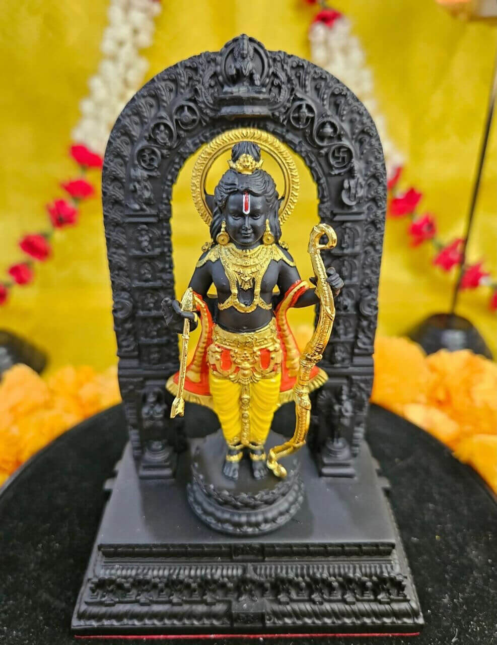 A detailed image of the black Ram Lalla idol from Ayodhya, depicting Lord Rama as a child adorned with golden jewelry, holding a golden bow and arrow, and placed within a black arch on a base, all against a vibrant yellow background.