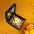 An open, dark wooden pooja box containing a golden Goddess Laxmi Charan Paduka (sacred footprints) set against a red fabric background with a decorative gold pattern. The box is placed on an orange surface with flower petals, next to a bowl of marigold flowers.