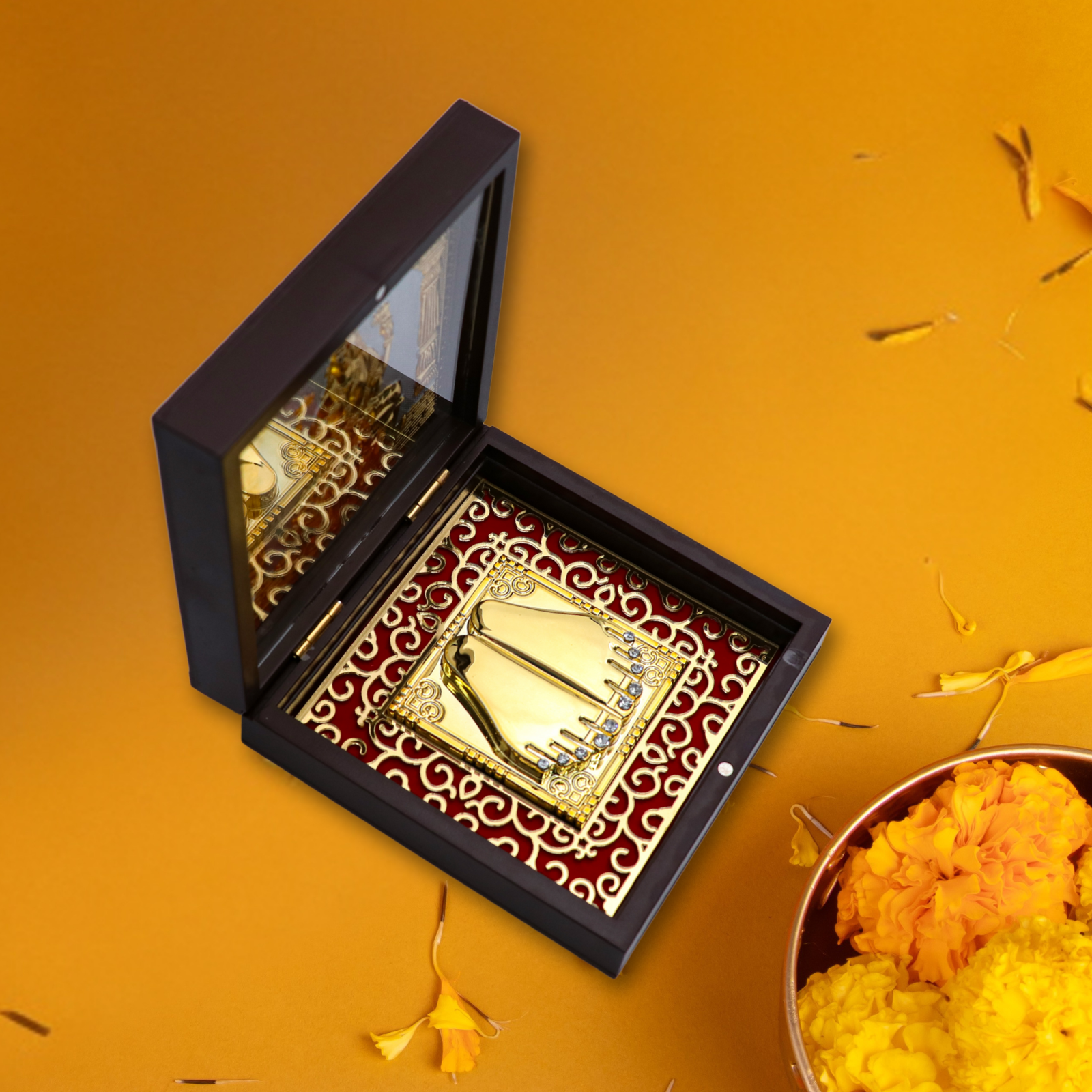 An open, dark wooden pooja box containing a golden Goddess Laxmi Charan Paduka (sacred footprints) set against a red fabric background with a decorative gold pattern. The box is placed on an orange surface with flower petals, next to a bowl of marigold flowers.