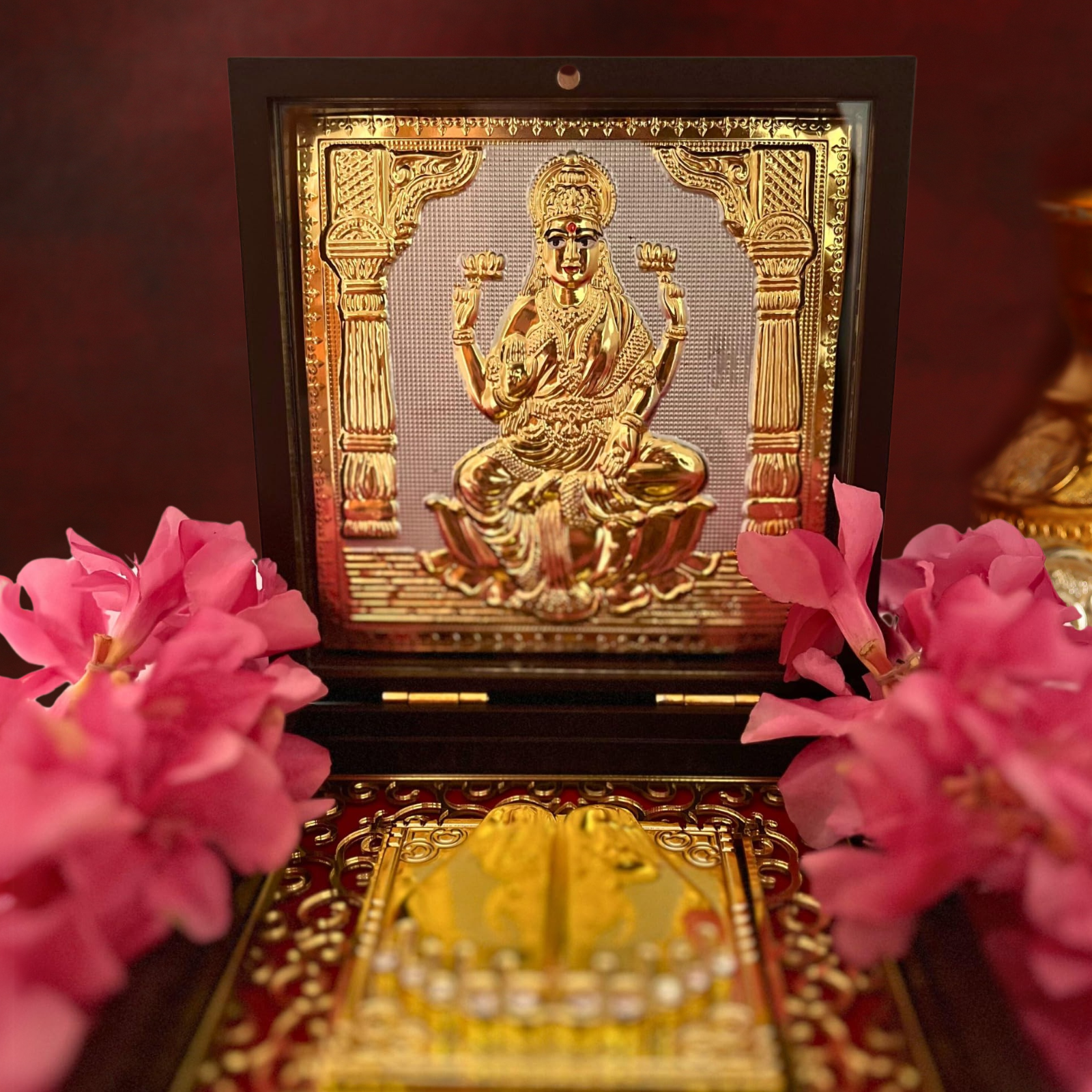 An open, dark wooden box containing a golden relief of Goddess Laxmi on the inside of the lid. The lower compartment contains a set of Golden Laxmi Charan Paduka (sacred footprints) set against a red, decorative background with gold patterns. The box is placed on a surface with pink flowers on either side.