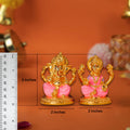 A close-up shot of a pair of small, golden-colored idols of the Hindu deities Ganesha and Lakshmi, placed side-by-side on a brown surface. Ganesha is on the left, sitting in a pink and gold throne-like base. Lakshmi is on the right, also seated on a matching pink and gold base. A ruler is visible on the left side of the image, indicating the Ganesha idol is 3 inches tall and both idols are 2 inches wide.
