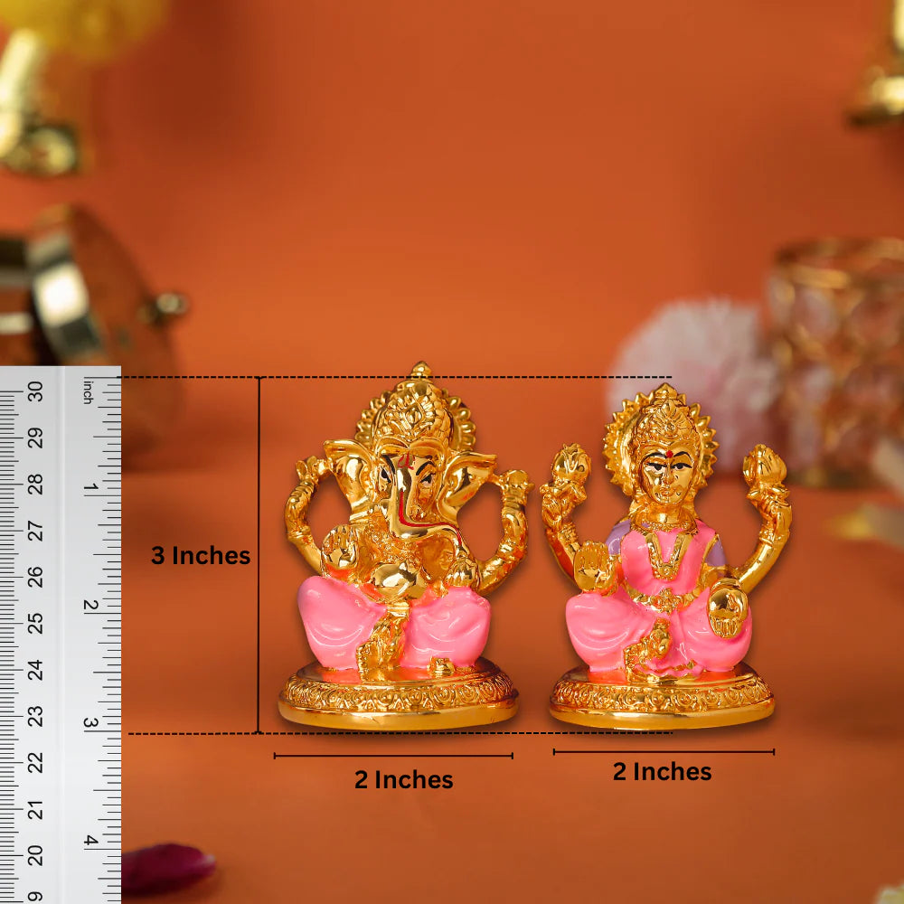A close-up shot of a pair of small, golden-colored idols of the Hindu deities Ganesha and Lakshmi, placed side-by-side on a brown surface. Ganesha is on the left, sitting in a pink and gold throne-like base. Lakshmi is on the right, also seated on a matching pink and gold base. A ruler is visible on the left side of the image, indicating the Ganesha idol is 3 inches tall and both idols are 2 inches wide.