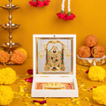 A decorative white box open to reveal a gold-plated idol of Lord Balaji (Venkateswara) and a Charan Paduka (footprints) on a patterned red and gold base. The box is surrounded by traditional Hindu puja items including lit brass lamps, yellow and orange marigold flowers, rose petals, and orange sweets (laddus) on a yellow surface.