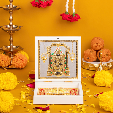 A decorative white box open to reveal a gold-plated idol of Lord Balaji (Venkateswara) and a Charan Paduka (footprints) on a patterned red and gold base. The box is surrounded by traditional Hindu puja items including lit brass lamps, yellow and orange marigold flowers, rose petals, and orange sweets (laddus) on a yellow surface.