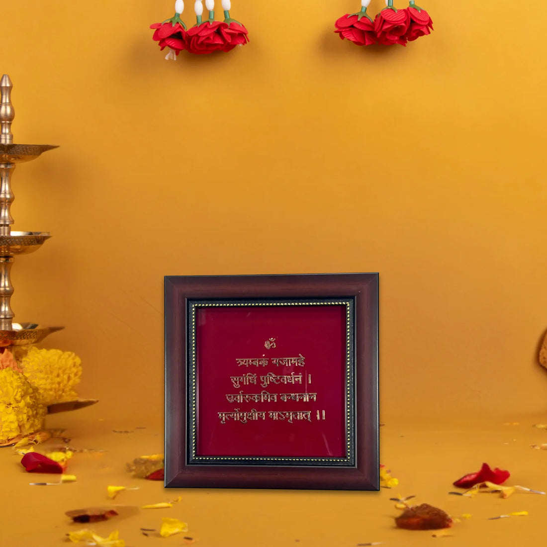 A wooden photo frame with a deep red background and golden text of - Mahamrityunjaya Mantra is placed on a surface. The text inside the frame is in Devanagari script. The frame is set against a yellow wall, with red flower garlands hanging above it. There are also some scattered flower petals and a brass lamp visible on the left side of the image.